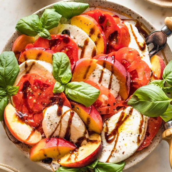 Earth-toned plate filled with a peach Caprese salad drizzled with balsamic glaze and fresh basil leaves.