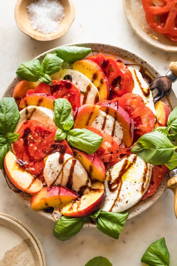 Earth-toned plate filled with a peach Caprese salad drizzled with balsamic glaze and fresh basil leaves.