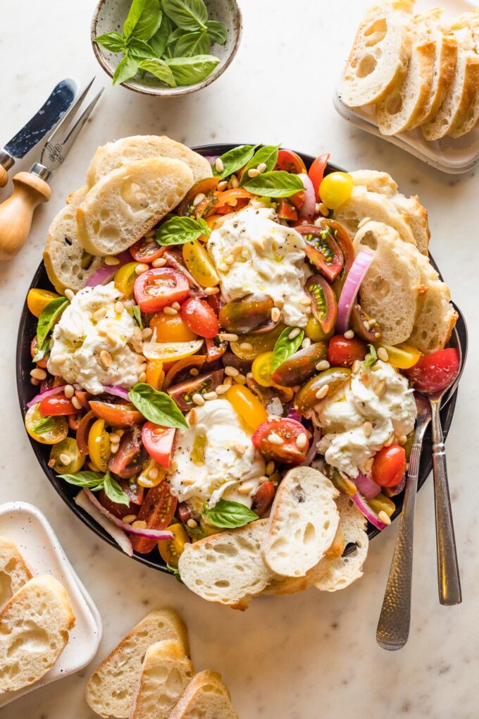 Platter with marinated tomatoes, burrata, pine nuts, and bread for serving.