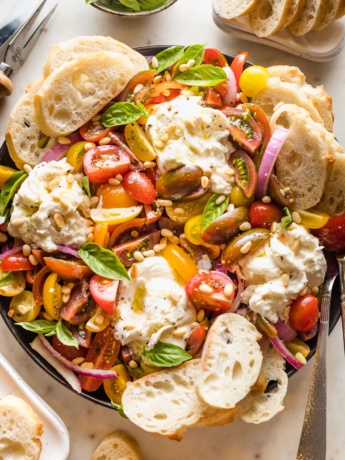 Platter with marinated tomatoes, burrata, pine nuts, and bread for serving.