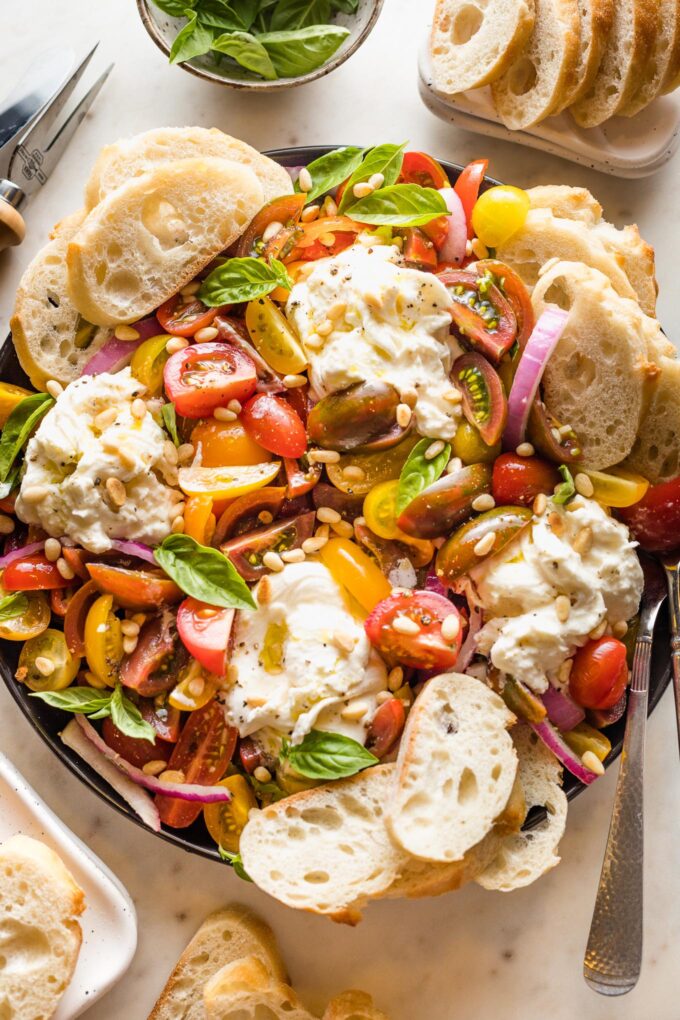 Platter with marinated tomatoes, burrata, pine nuts, and bread for serving.