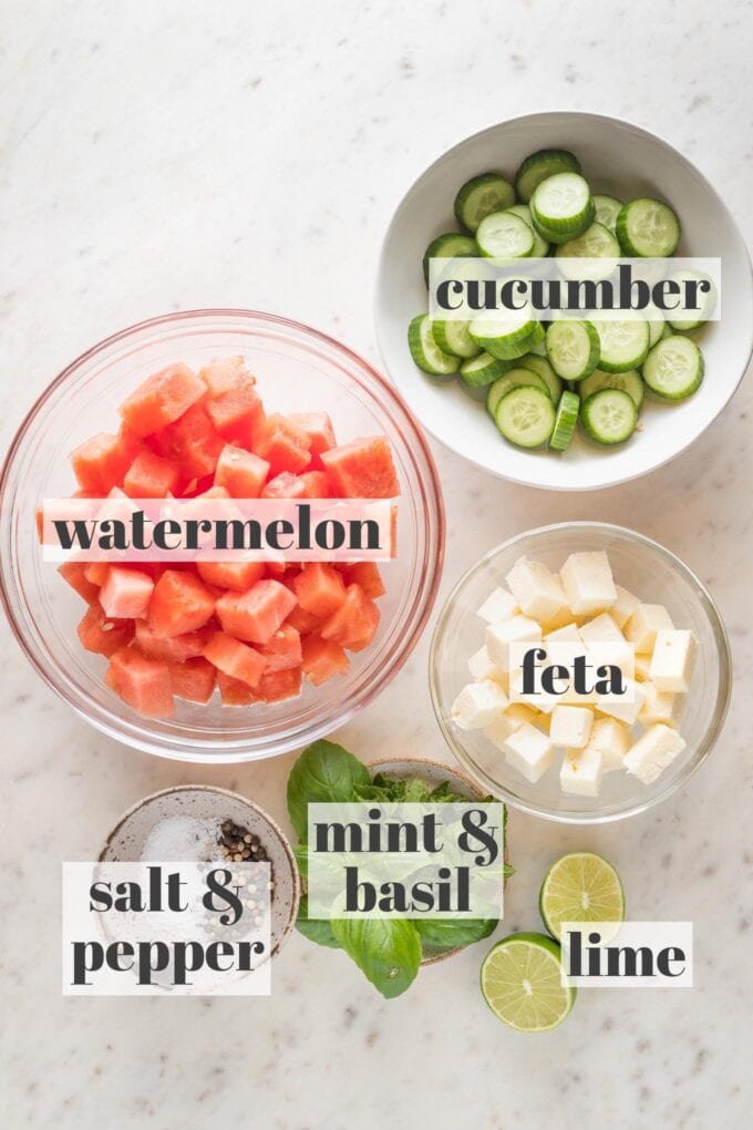 Prep bowls holding cut watermelon, baby cucumbers, feta cheese, fresh mint, fresh basil, limes, salt, and pepper.