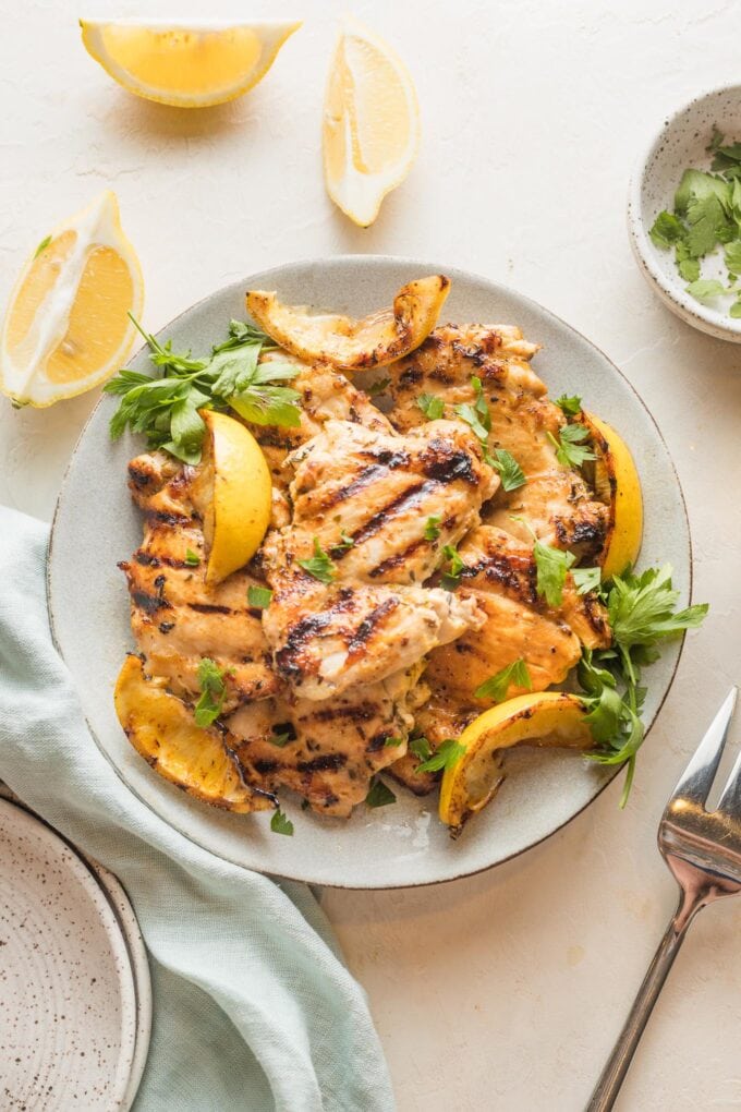 Small plate with lemon chicken thighs made in a grill pan surrounded by parsley and lemon wedges.