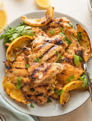 Plate piled with lemon chicken thighs served with parsley and grilled lemon wedges.