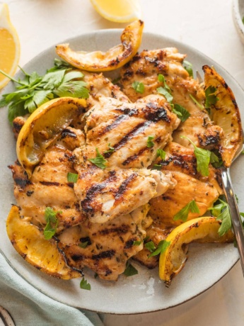 Plate piled with lemon chicken thighs served with parsley and grilled lemon wedges.