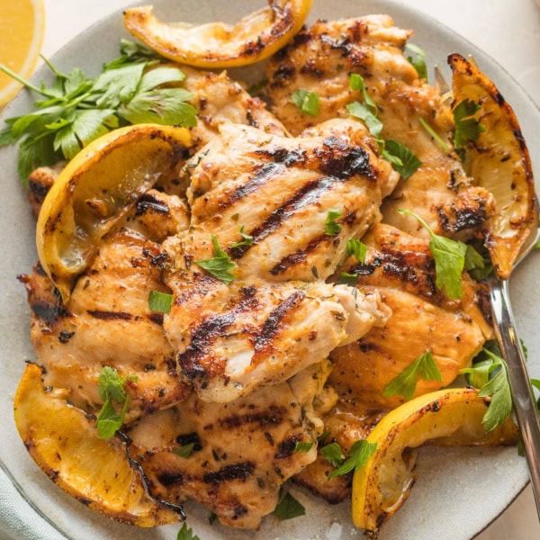 Plate piled with lemon chicken thighs served with parsley and grilled lemon wedges.