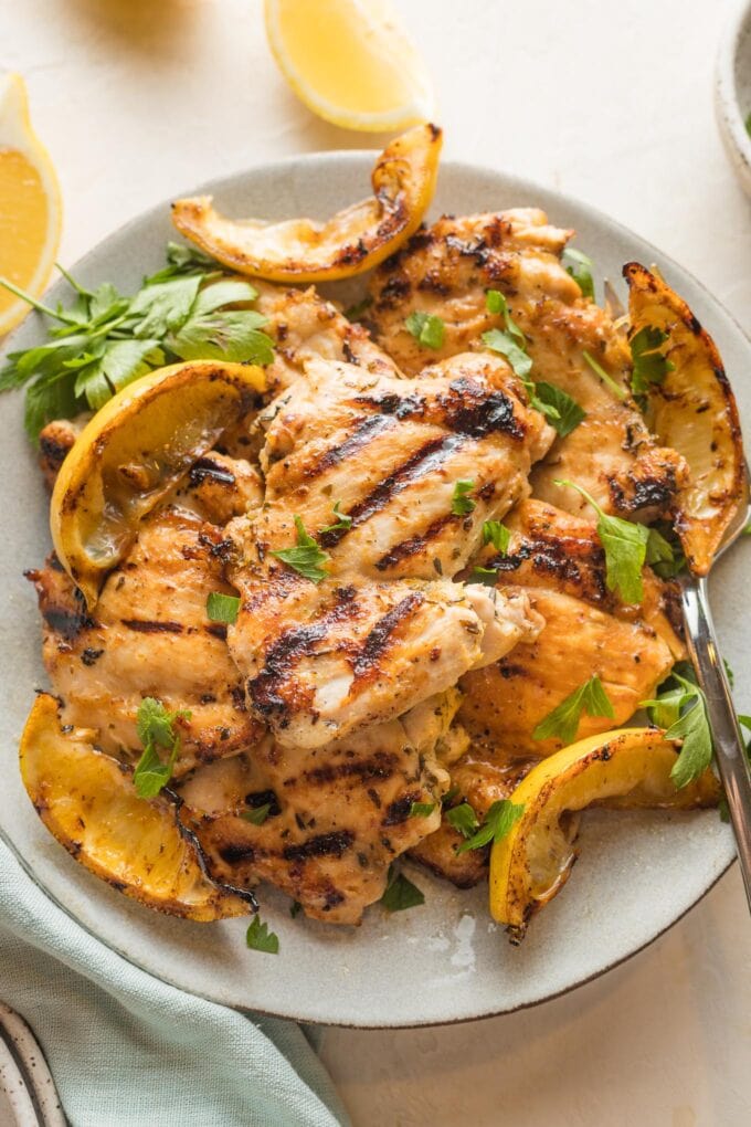Plate piled with lemon chicken thighs served with parsley and grilled lemon wedges.