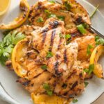 Plate piled with lemon chicken thighs served with parsley and grilled lemon wedges.