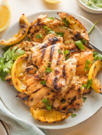 Plate piled with lemon chicken thighs served with parsley and grilled lemon wedges.