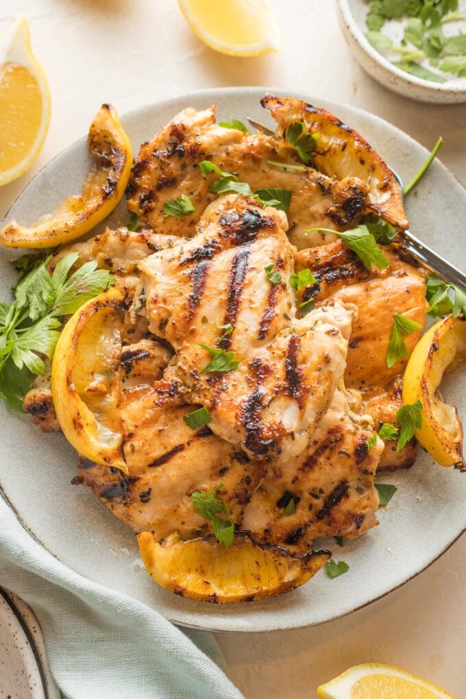 Plate piled with lemon chicken thighs served with parsley and grilled lemon wedges.