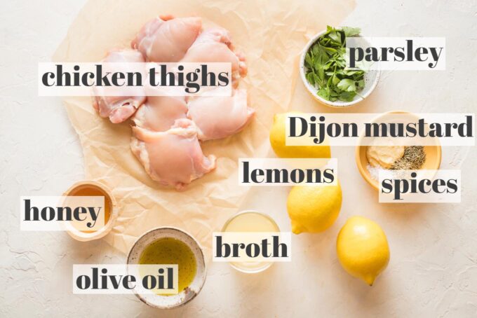 Raw chicken thighs on parchment paper surrounded by prep bowls holding honey, olive oil, chicken broth, lemons, Dijon mustard, parsley, and spices.