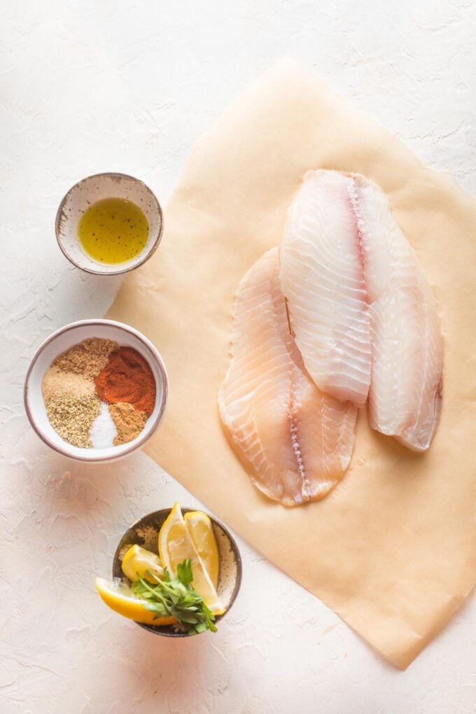 Overhead image of tilapia fillets, olive oil, spices, and lemon wedges.