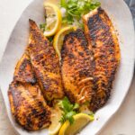 Platter with blackened tilapia fillets served with lemon wedges and a garnish of fresh parsley.