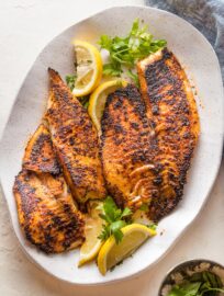 Platter with blackened tilapia fillets served with lemon wedges and a garnish of fresh parsley.