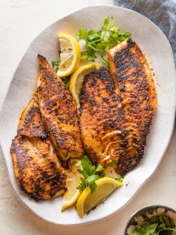 Platter with blackened tilapia fillets served with lemon wedges and a garnish of fresh parsley.