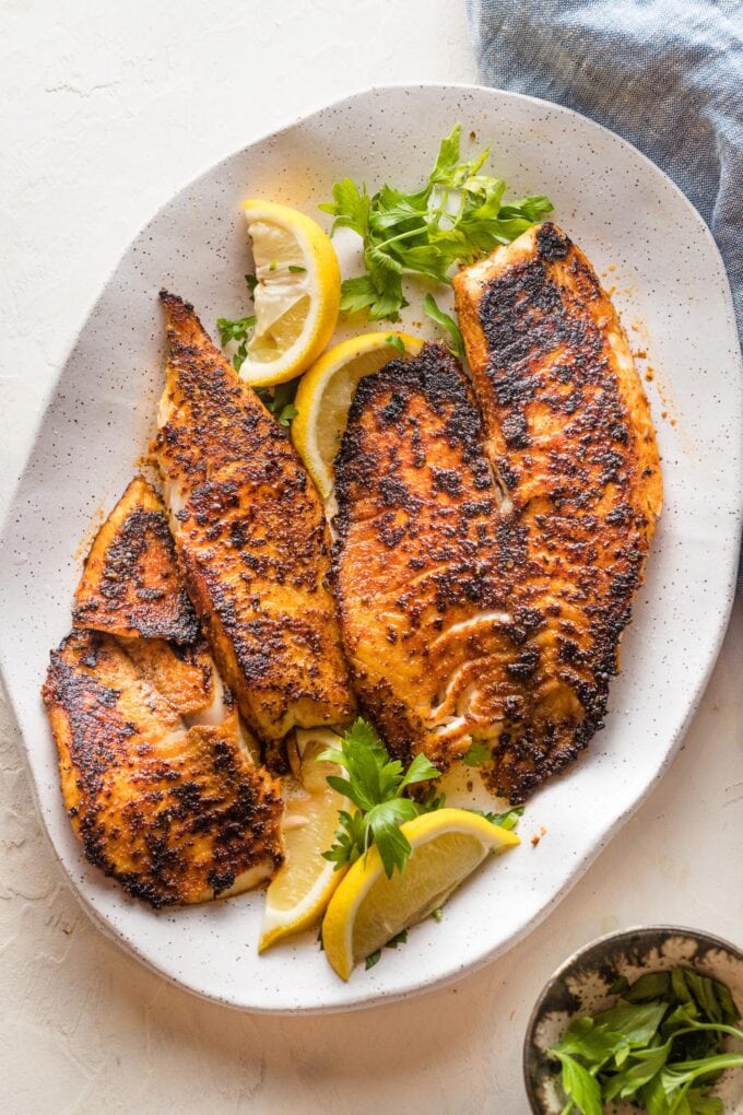 Platter with blackened tilapia fillets served with lemon wedges and a garnish of fresh parsley.