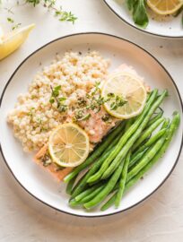 White plate with a serving of baked lemon herb salmon garnished with fresh thyme, served with green beans and pearl couscous.