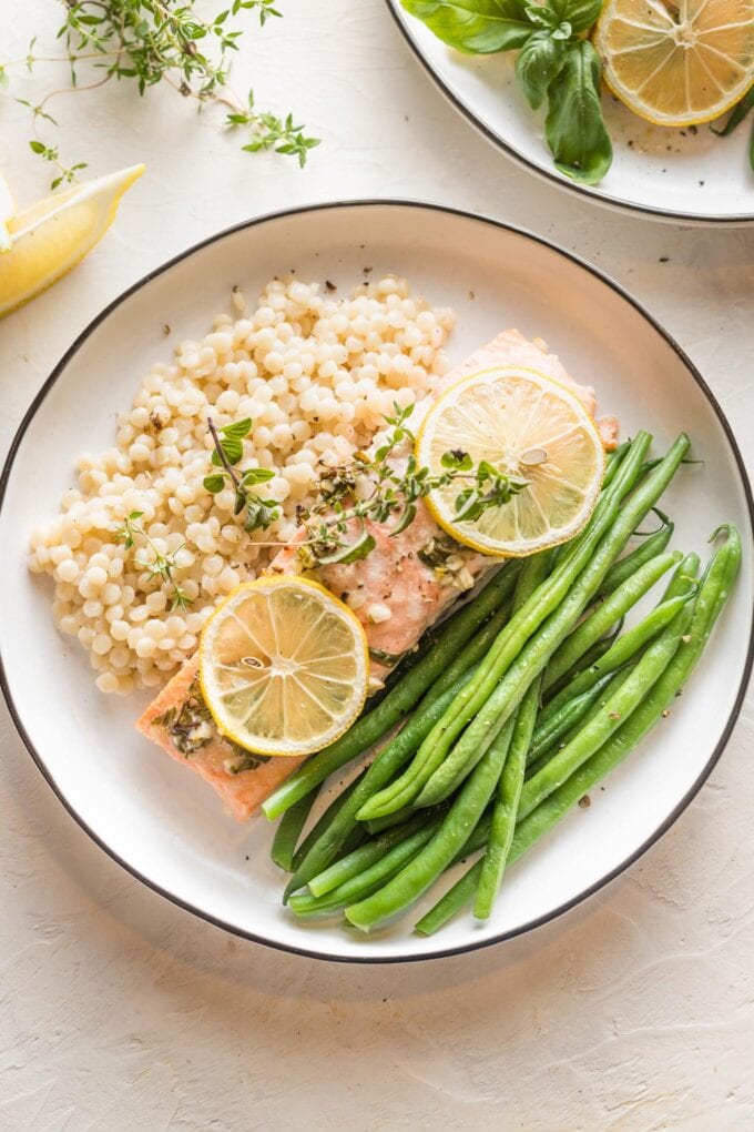 White plate with a serving of baked lemon herb salmon garnished with fresh thyme, served with green beans and pearl couscous.