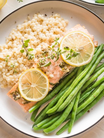 White plate with a serving of baked lemon herb salmon garnished with fresh thyme, served with green beans and pearl couscous.