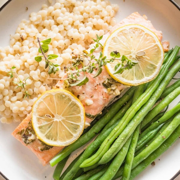 White plate with a serving of baked lemon herb salmon garnished with fresh thyme, served with green beans and pearl couscous.