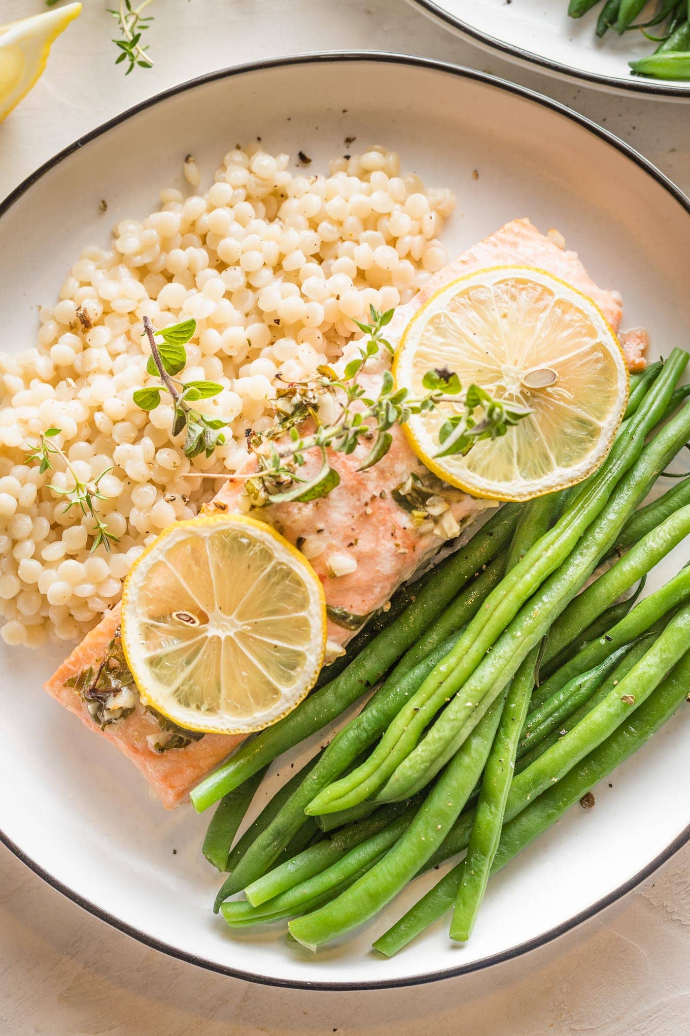White plate with a serving of baked lemon herb salmon garnished with fresh thyme, served with green beans and pearl couscous.
