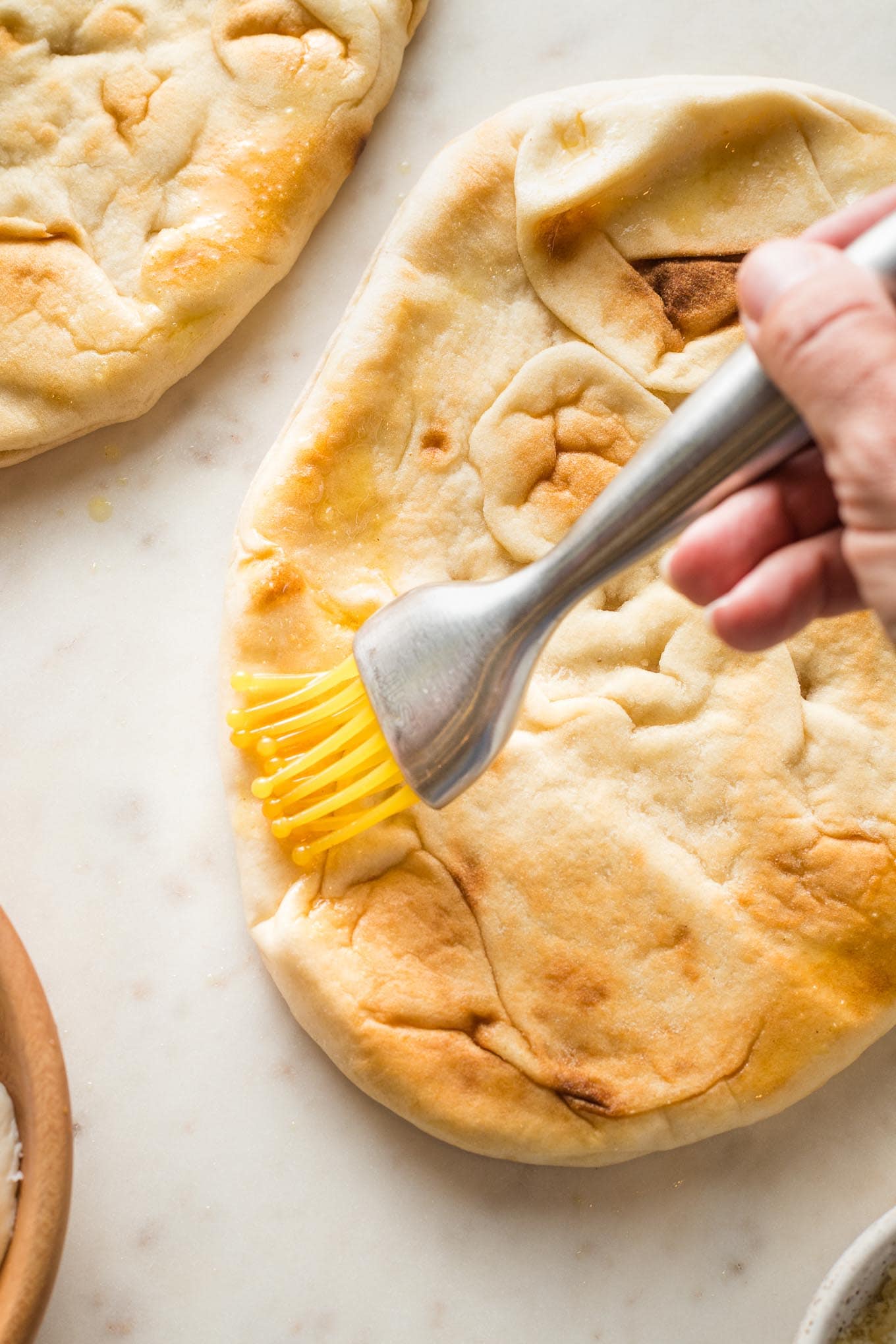 A small pastry brush spreading olive oil around the outer edge of a naan.