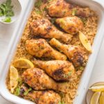 Large rectangular baking pan filled with baked chicken legs, rice, herbs, and lemon wedges.