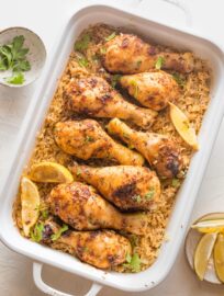 Large rectangular baking pan filled with baked chicken legs, rice, herbs, and lemon wedges.