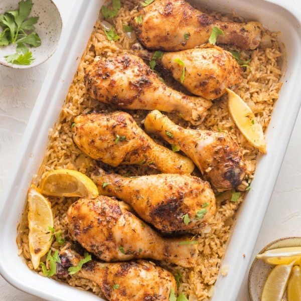 Large rectangular baking pan filled with baked chicken legs, rice, herbs, and lemon wedges.