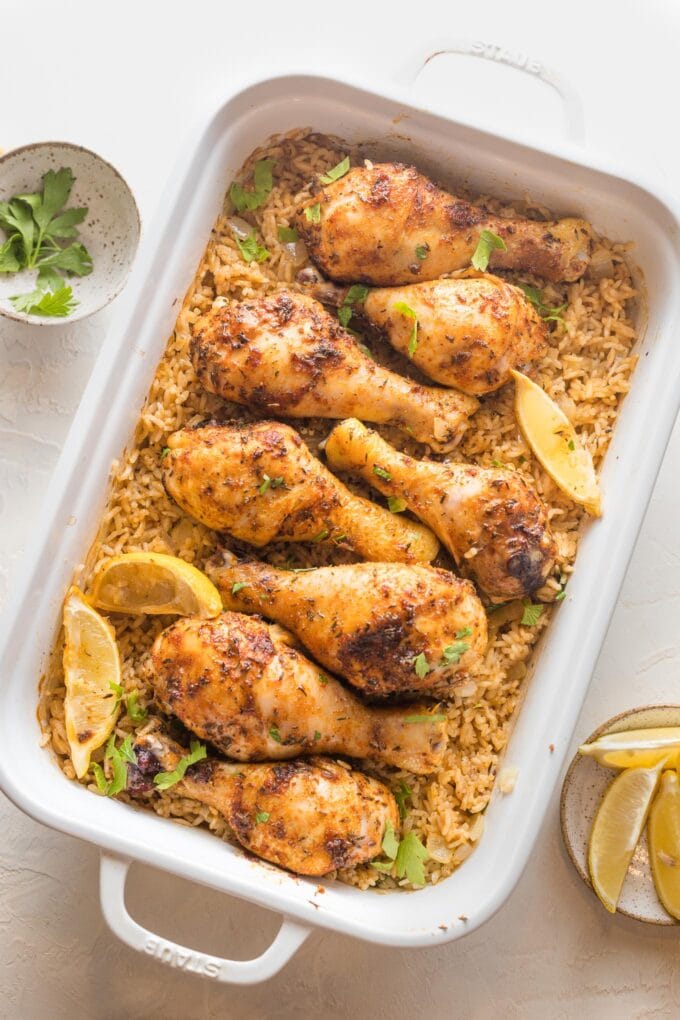 Large rectangular baking pan filled with baked chicken legs, rice, herbs, and lemon wedges.