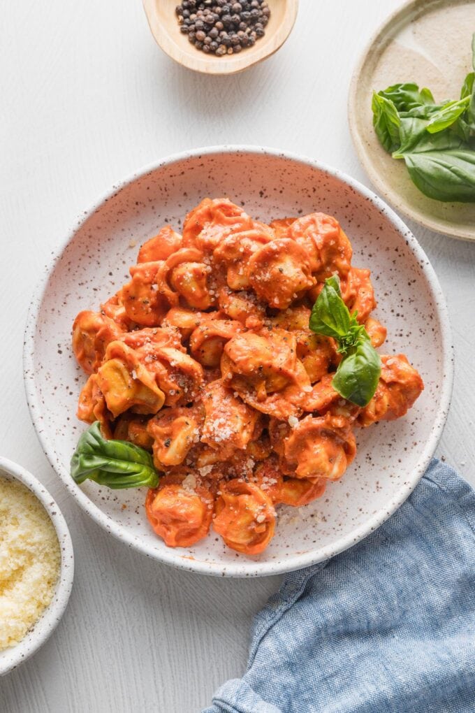Table set with bowls of creamy tomato tortellini, with fresh basil, grated Parmesan, and black pepper in a grinder for extra flavor.