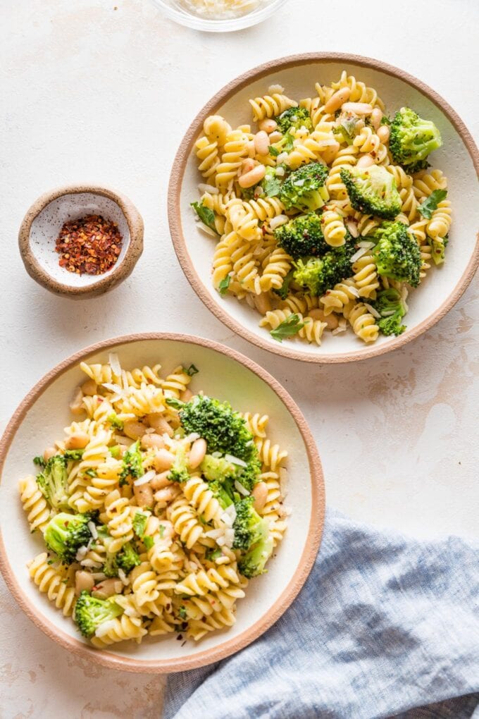 Two bowls of pasta with white beans and broccoli with a small dish of red pepper flakes for garnish.