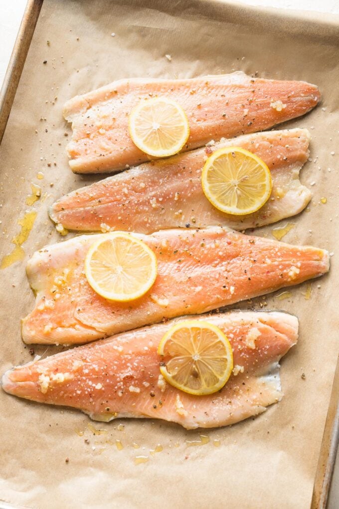 Uncooked rainbow trout filets on a sheet pan with garlic and lemon on top.