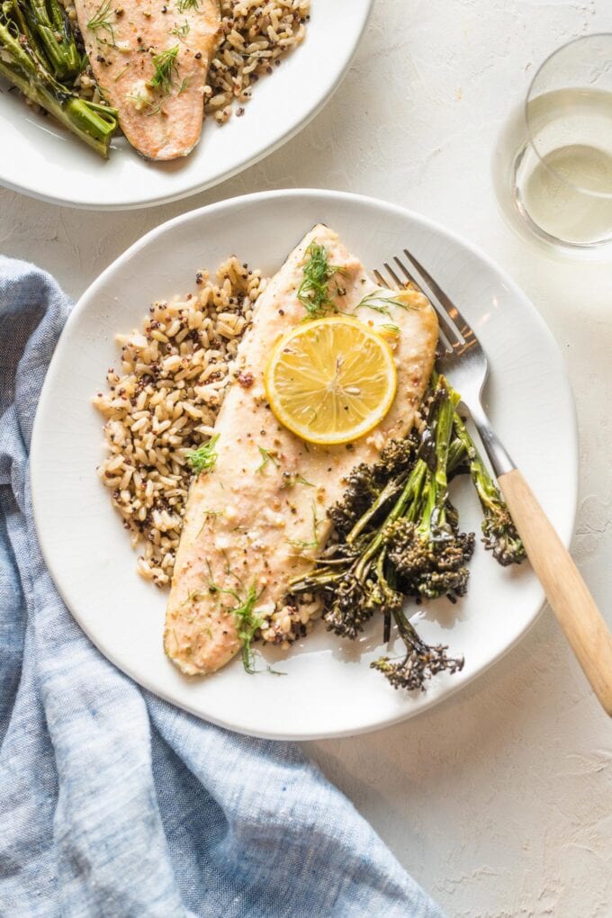Rainbow trout baked with lemon and garlic and served on a small white plate along with roasted broccolini and a wild and brown rice mix.