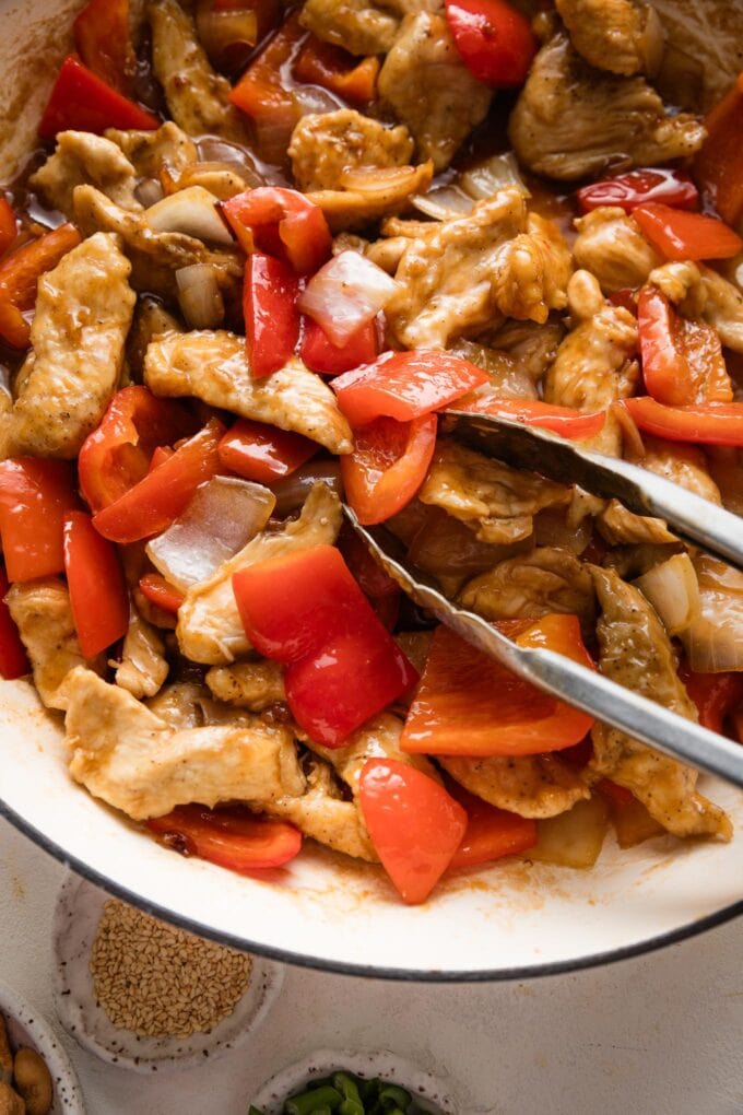 Close up of kitchen tongs picking up pieces of stir fry chicken and peppers.