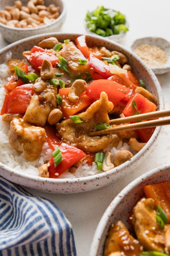 Close up of chopsticks picking up a piece of thin-sliced stir fry chicken from a bowl with peppers, onions, cashews, and homemade sauce.