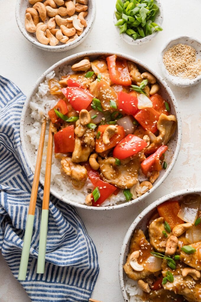 Countertop set with two bowls full of a homemade chicken and bell pepper stir fry with extra cashews, green onions, and sesame seeds nearby as toppings.