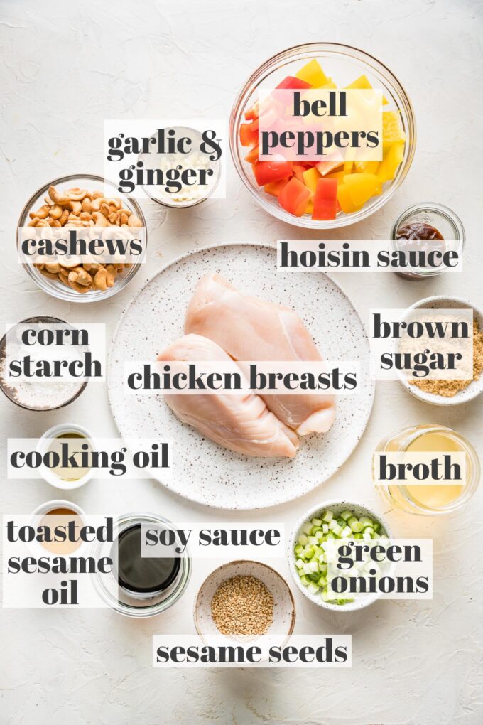 Labeled photo with chicken breasts, bell peppers, garlic, ginger, cashews, corn starch, cooking oil, toasted sesame oil, soy sauce, sesame seeds, green onions, broth, brown sugar, and hoisin sauce all in prep bowls and on plates.