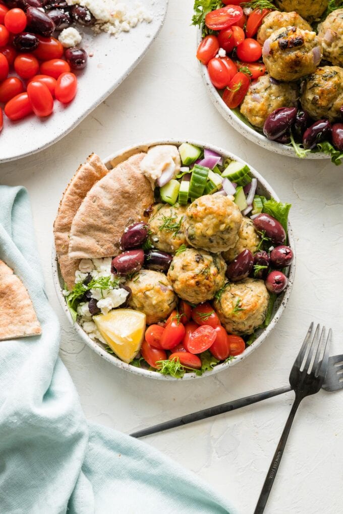 Two Greek bowls with chicken meatballs, olives, veggies, pita bread, and hummus.