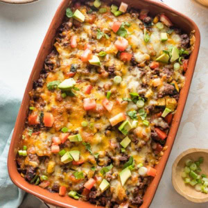 Enchilada casserole with ground beef, veggies, and chopped avocado for toppings, served in a brown enameled baking pan.