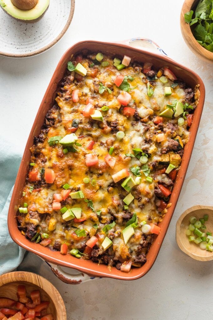 Enchilada casserole with ground beef, veggies, and chopped avocado for toppings, served in a brown enameled baking pan.