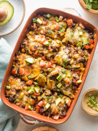 Enchilada casserole with ground beef, veggies, and chopped avocado for toppings, served in a brown enameled baking pan.