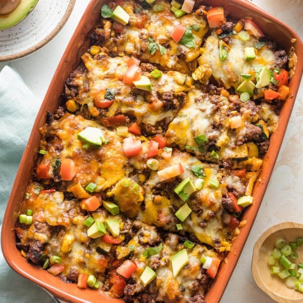 Enchilada casserole with ground beef, veggies, and chopped avocado for toppings, served in a brown enameled baking pan.