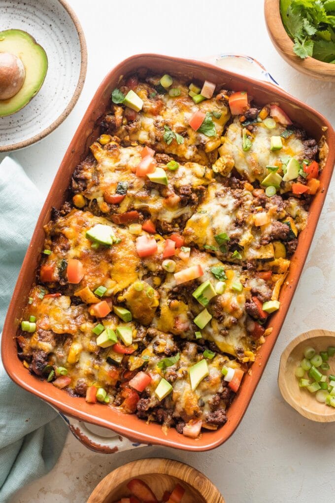 Enchilada casserole with ground beef, veggies, and chopped avocado for toppings, served in a brown enameled baking pan.