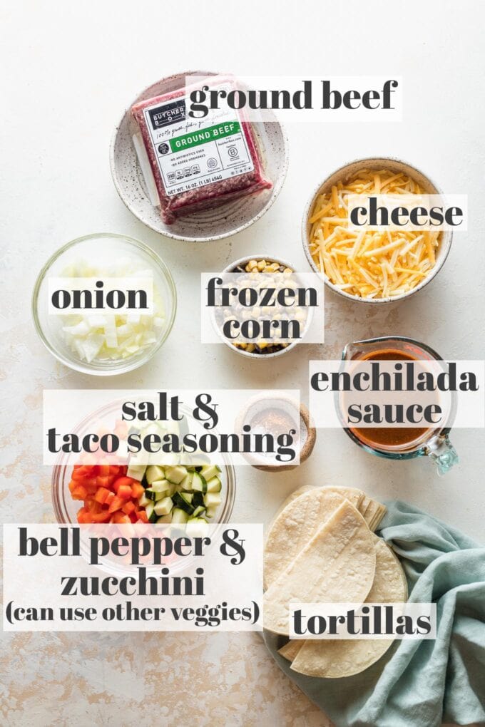 Labeled photo of ground beef, shredded Mexican cheese, onion, frozen corn, enchilada sauce, salt, taco seasoning, chopped zucchini and bell pepper, and tortillas in prep bowls.