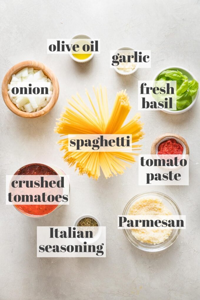 Labeled photo with spaghetti, crushed tomatoes, fresh basil, tomato paste, Parmesan, Italian seasoning, garlic, onion, and olive oil in prep bowls.