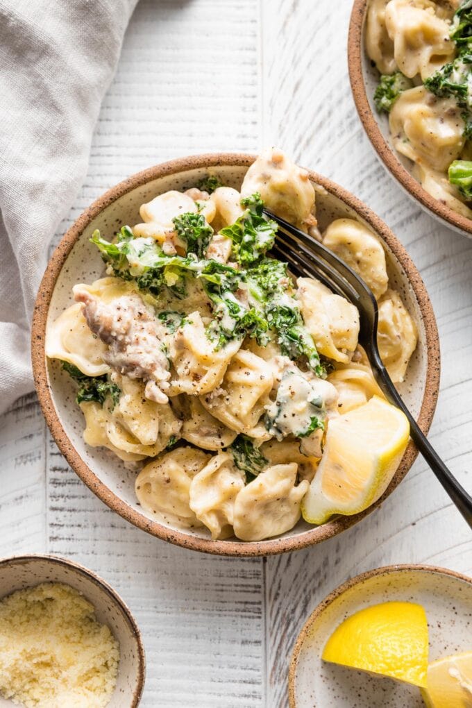 Small serving bowl with sausage tortellini Alfredo served with kale and a squeeze of lemon juice.