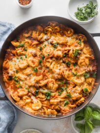 Countertop with a large, deep skillet of baked tortellini with meat sauce, with extra herbs and red pepper flakes nearby for sprinkling.