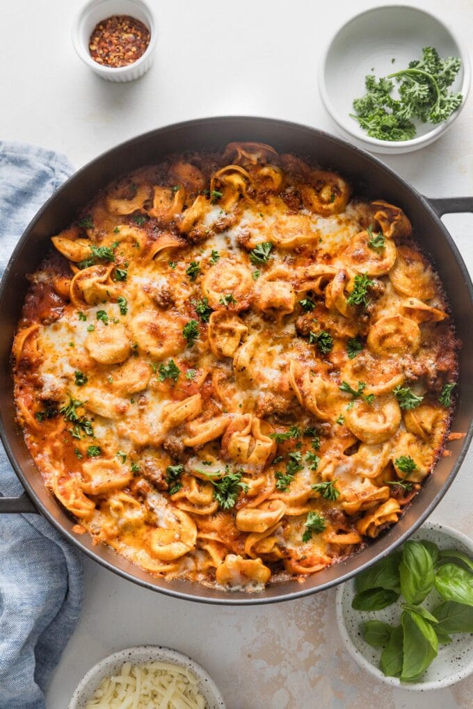 Countertop with a large, deep skillet of baked tortellini with meat sauce, with extra herbs and red pepper flakes nearby for sprinkling.