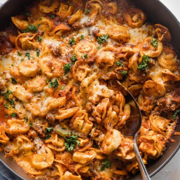 Large cast iron skillet full of baked tortellini with a spoon poised to serve.